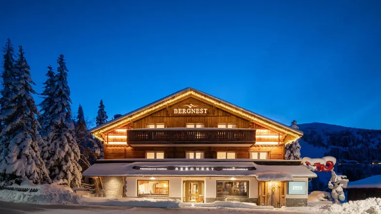 Beleuchtetes Holzgebäude in einer verschneiten Winternacht, umgeben von Bäumen und Bergen.