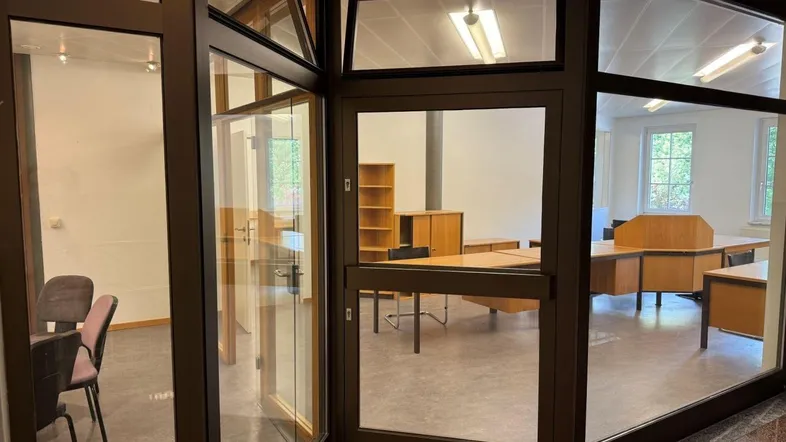 Blick durch eine Glaswand in ein helles Büro mit mehreren Schreibtischen und Holzmöbeln.