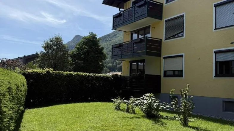 Gelbe Fassade eines Mehrfamilienhauses mit Balkonen und gepflegtem Rasen im Vordergrund, umgeben von Bergen.