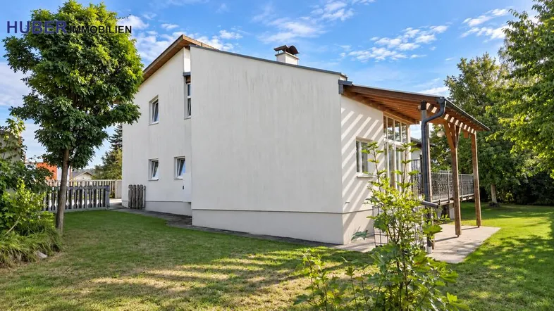 Hausfassade mit großem Gartenbereich, Rasenfläche und einer überdachten Terrasse mit Pergola.