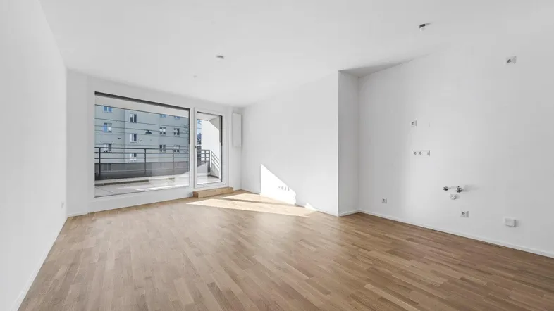 Helles, leeres Wohnzimmer mit Parkettboden und großen Fenstern, die Zugang zu einem Balkon bieten.