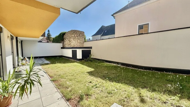 Sonniger Gartenbereich mit Rasenfläche, gefliester Terrasse und dekorativer Steinmauer.