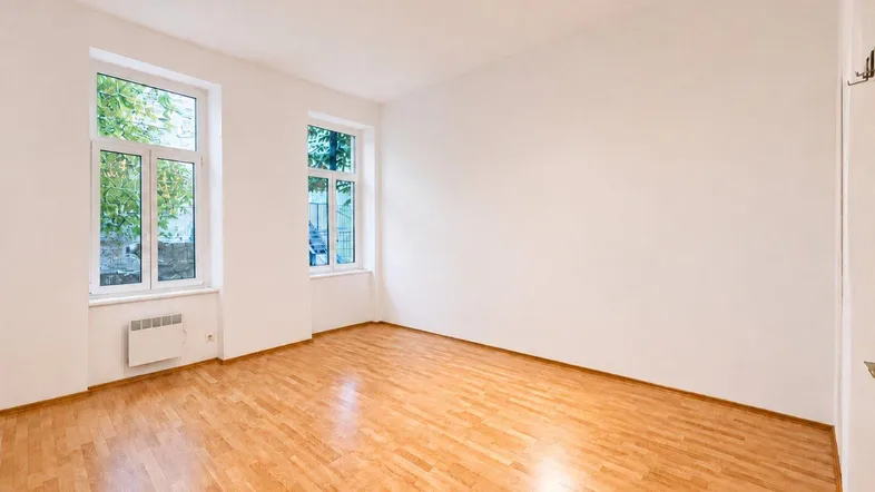 Helles, leeres Zimmer mit Parkettboden und zwei großen Fenstern, ideal als Wohn- oder Schlafzimmer.