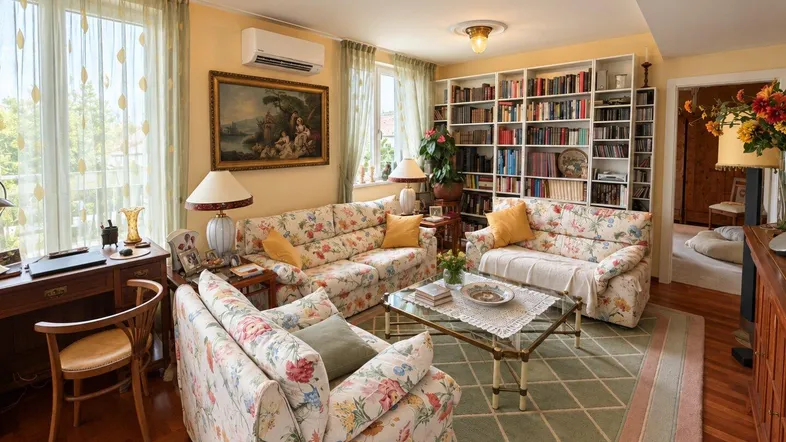 Gemütliches Wohnzimmer mit floralen Sofas, großem Bücherregal und Zugang zu einem sonnigen Balkon.