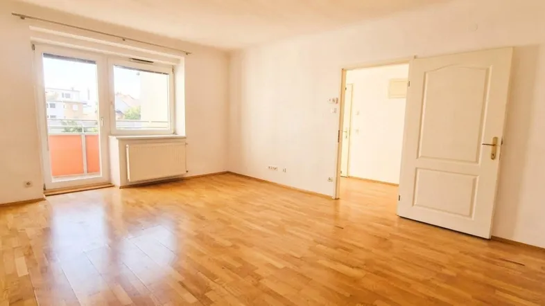 Helles Zimmer mit Parkettboden, weißen Wänden und Zugang zu einem kleinen Balkon.