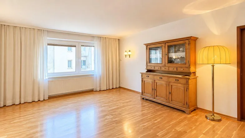 Helles, leeres Wohnzimmer mit Holzboden, großem Fenster und traditionellem Holzschrank.
