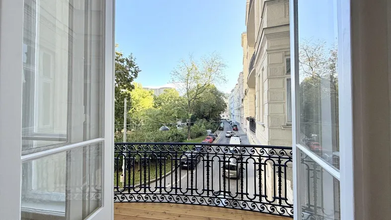 Geräumiger Balkon mit Holzboden und schmiedeeisernem Geländer, bietet Blick auf die Straße und Bäume.