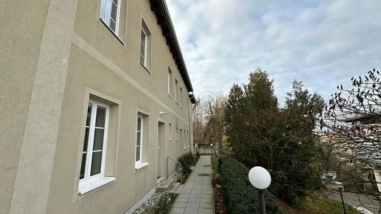 Die Fassade des Gebäudes mit Fenstern und einem gepflasterten Weg, umgeben von Bäumen.