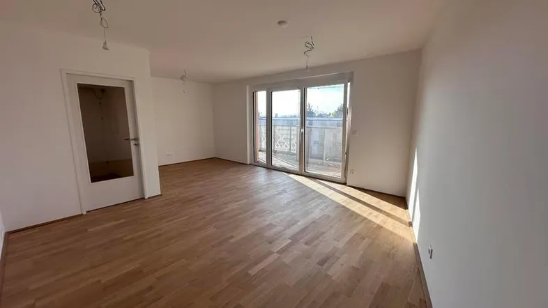 Helles, leeres Wohnzimmer mit Parkettboden und Zugang zu einem sonnigen Balkon.