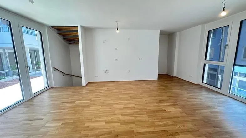 Helles, leeres Wohnzimmer mit Parkettboden, großen Fenstern und Zugang zu einem Außenbereich.
