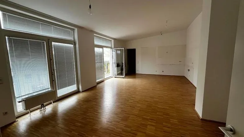 Geräumiger, leerer Wohnbereich mit Parkettboden, großen Fenstern und Zugang zu einem Balkon.