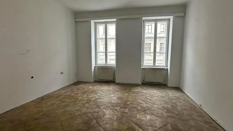 Helles, leeres Zimmer mit Fischgrätparkett und zwei Fenstern, bereit zur Renovierung.