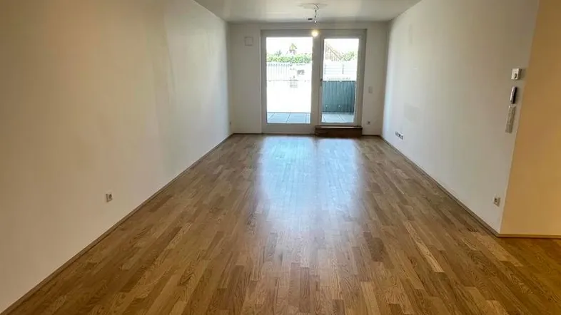 Heller, leerer Raum mit Holzboden und Zugang zu einem Außenbereich durch eine Glastür.
