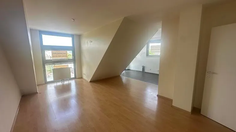 Helles Zimmer mit Dachschrägen, Holzboden und großen Fenstern, das in einen weiteren Raum übergeht.