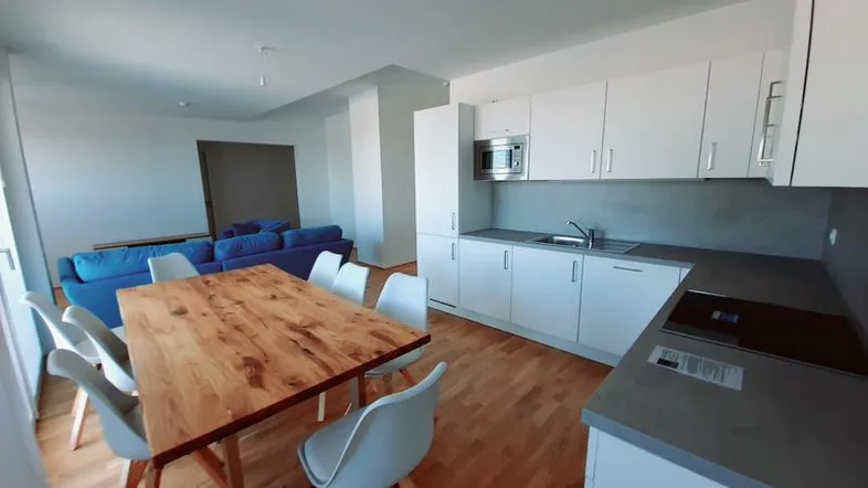 Heller offener Wohn-Essbereich mit moderner Einbauküche, Holztisch und blauem Sofa.