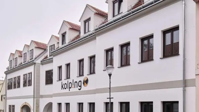 Modernes Gebäude mit weißer Fassade, Satteldach und mehreren Gaubenfenstern an einem bewölkten Tag.