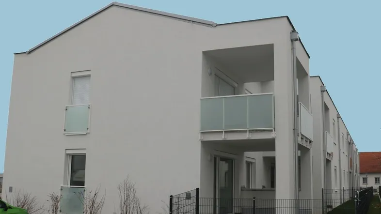 Modernes Mehrfamilienhaus mit weißer Fassade und verglasten Balkonen, umgeben von einer kleinen Grünfläche.