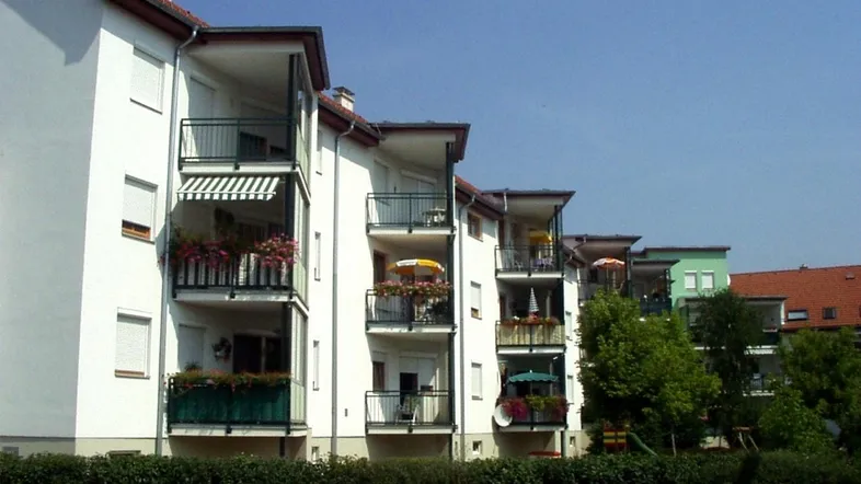 Mehrfamilienhaus mit weißer Fassade, zahlreichen Balkonen und grüner Umgebung mit Spielplatz.