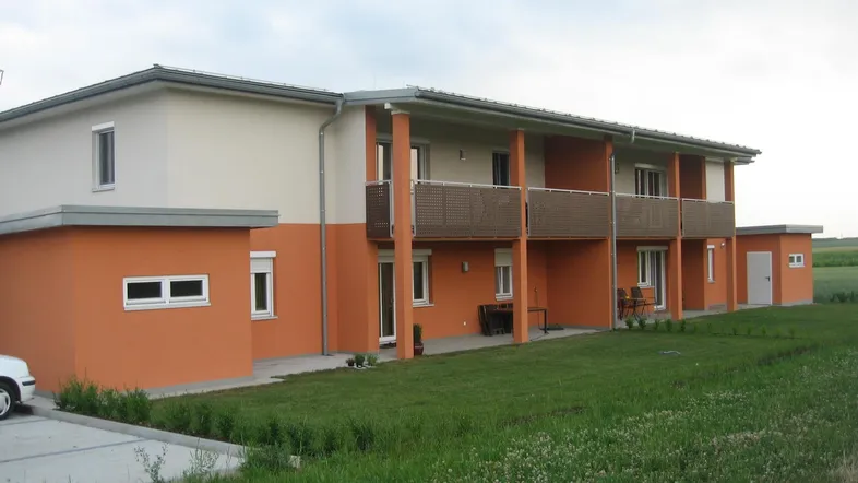 Modernes Mehrfamilienhaus mit orangefarbener Fassade, Balkonen und Grünflächen in ländlicher Umgebung.