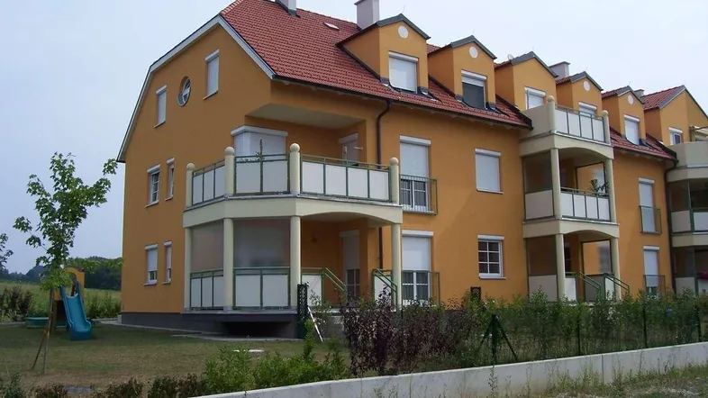Mehrfamilienhaus mit gelber Fassade, roten Ziegeldächern und Balkonen, umgeben von Grünflächen und einem kleinen Spielplatz.