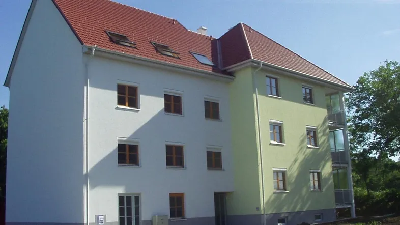 Neues Mehrfamilienhaus mit weißer und hellgrüner Fassade, rotem Satteldach und verglasten Balkonen.
