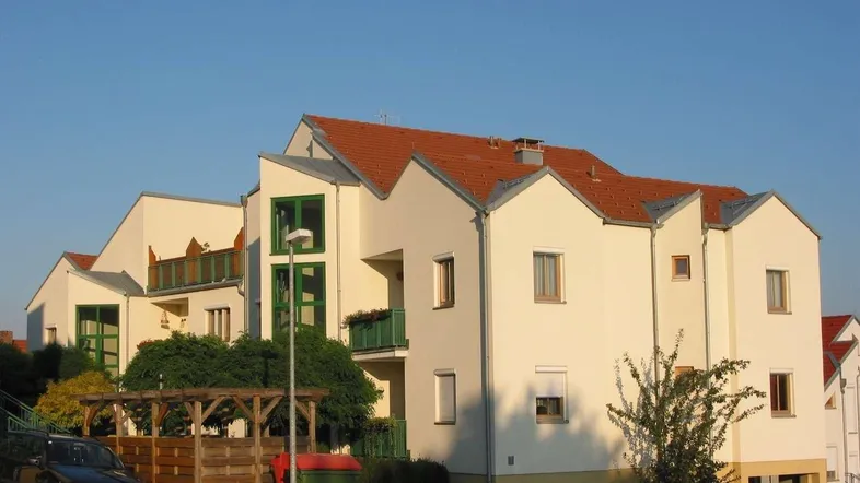 Modernes Mehrfamilienhaus mit heller Fassade, roten Ziegeldächern und grünen Fensterrahmen in sonniger Lage.