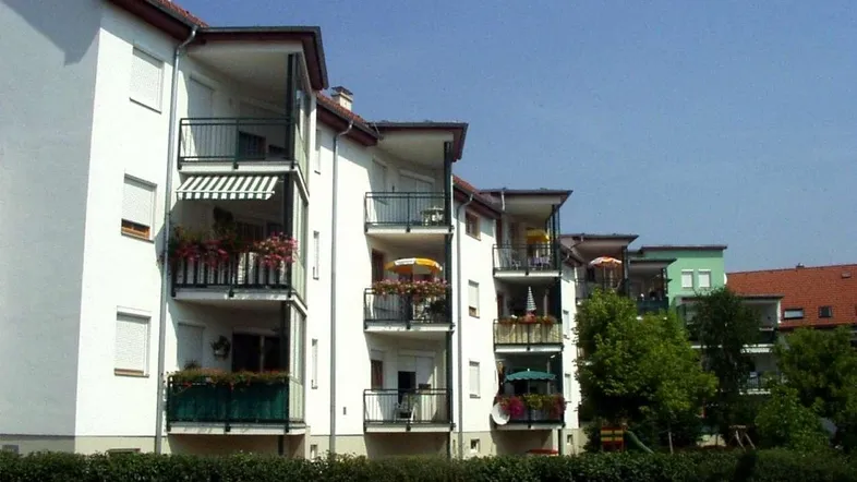 Mehrfamilienhaus mit mehreren Balkonen, umgeben von einer Hecke und einem kleinen Spielplatz.