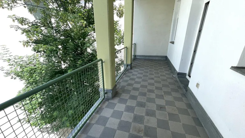 Geräumiger Balkon mit karierten Fliesen und grünem Gittergeländer, umgeben von Bäumen.