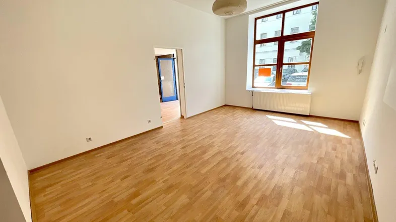 Großer heller Raum mit Holzboden und einem hohen Fenster, das viel Tageslicht hereinlässt.