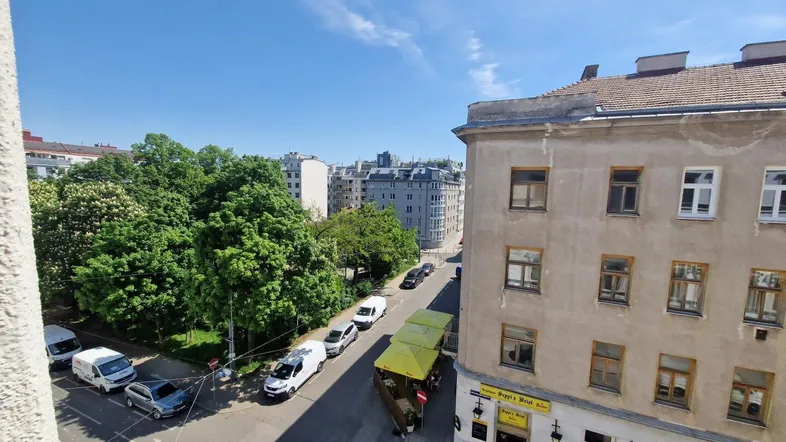 Blick von einem oberen Stockwerk auf eine belebte Straße mit Bäumen und gegenüberliegenden Gebäuden.