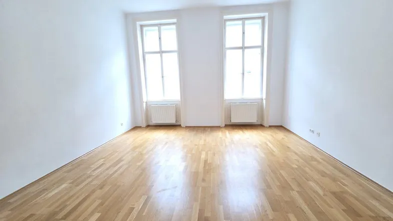 Helles Zimmer mit Holzboden und zwei großen Fenstern, ideal als Wohn- oder Schlafzimmer nutzbar.
