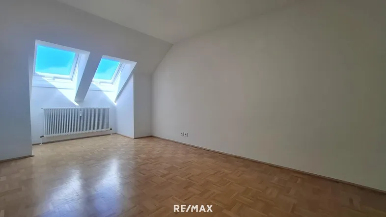 Helles Dachzimmer mit Parkettboden, weißen Wänden und zwei Dachfenstern, die viel Licht hereinlassen.