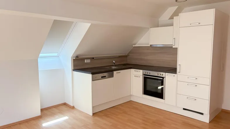 Moderne Einbauküche in einem Dachgeschossraum mit weißen Schränken, dunkler Arbeitsplatte und Dachfenster.