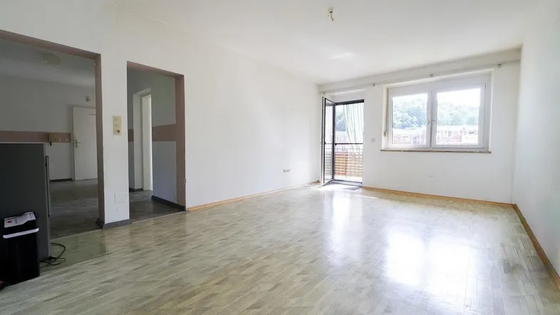 Helles, leeres Wohnzimmer mit Parkettboden und Zugang zum Balkon.