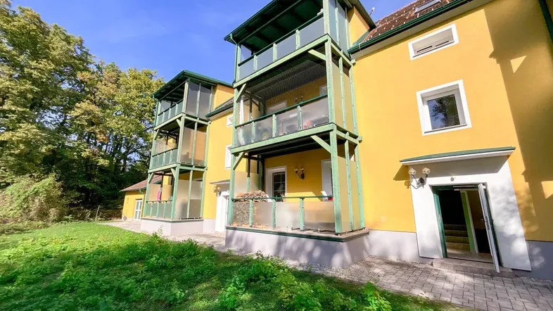 Gelbe Fassade eines Mehrfamilienhauses mit grünen Balkonen und umgebender Natur.