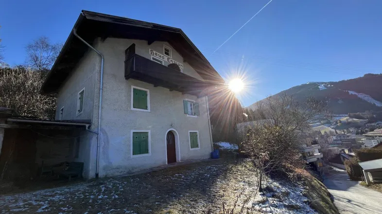 Traditionelles Haus mit Balkon und Satteldach in alpiner Umgebung unter strahlendem Sonnenschein.