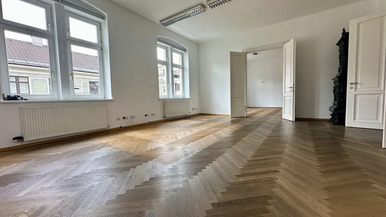 Geräumiger, heller Raum mit Fischgrätparkett, hohen Fenstern und Blick in einen weiteren Raum.