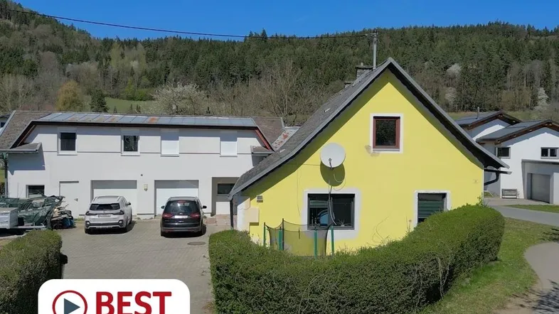 Gelbes Haus mit angrenzender Garage und Stellplätzen, umgeben von grüner Landschaft.