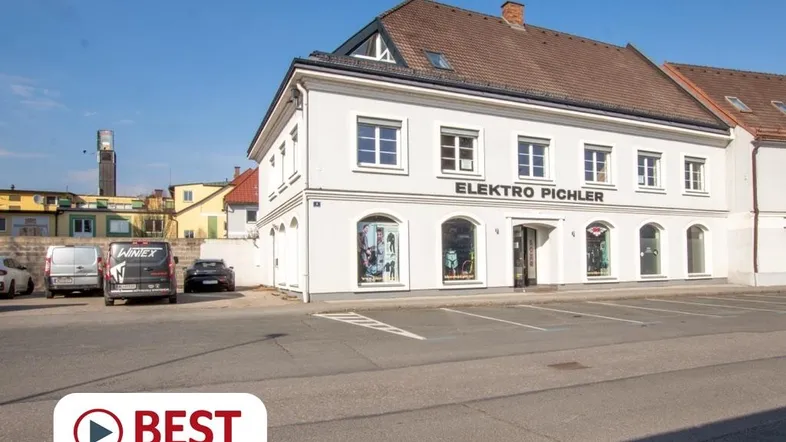 Weißes Geschäftsgebäude mit großen Schaufenstern und Parkplätzen davor unter blauem Himmel.