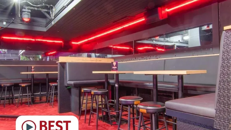Innenansicht einer Bar mit roten Neonlichtern, gepolsterten Sitzbänken und Hockern an Holztischen.