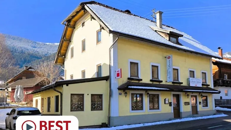 Gelbes Gebäude mit traditioneller Architektur, schneebedecktem Dach und Bergkulisse im Hintergrund.