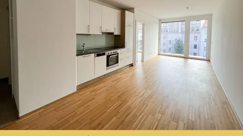Heller offener Wohnbereich mit integrierter Küche, Parkettboden und Zugang zum Balkon.