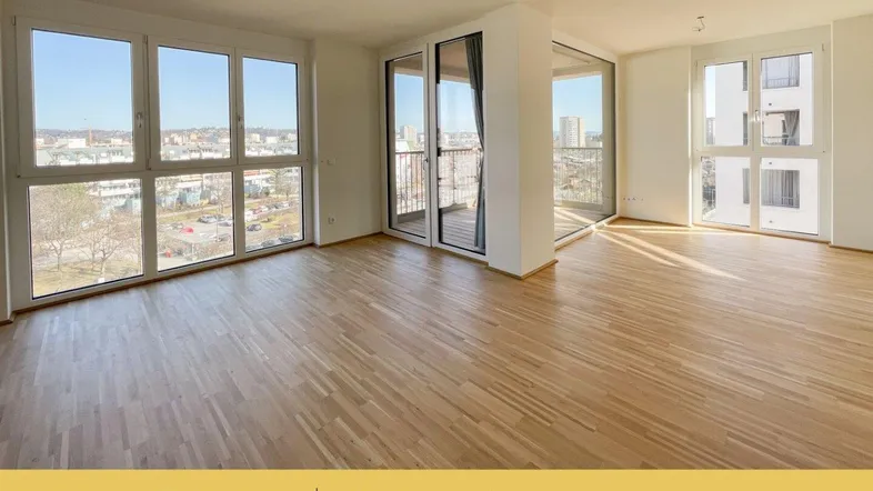 Helles, leeres Wohnzimmer mit Parkettboden, großen Fenstern und Zugang zum Balkon mit Stadtblick.