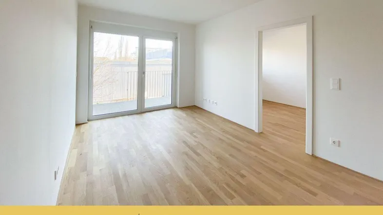 Helles, leeres Zimmer mit Holzboden und Zugang zu einem Balkon, ideal als Wohnzimmer.