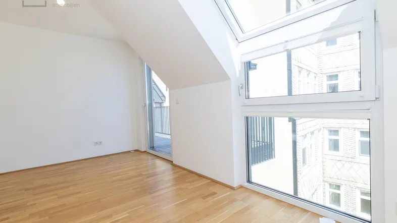 Helles, leeres Zimmer mit Parkettboden, großen Fenstern und Zugang zu einem Balkon.