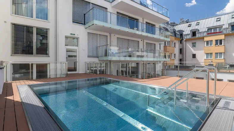 Moderner Außenpool auf einer Holzterrasse, umgeben von einem Gebäude mit Glasbalkonen.
