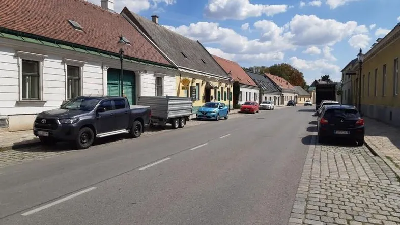 Straße mit traditionellen Gebäuden, geparkten Autos und Kopfsteinpflaster unter blauem Himmel.
