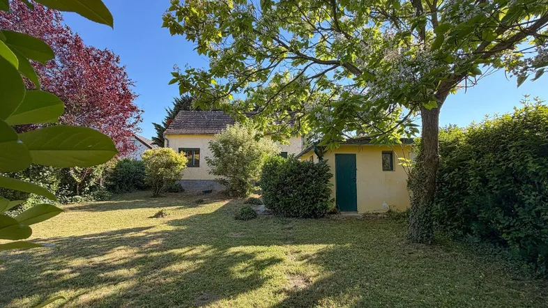 Gartenbereich mit Rasenfläche und Bäumen vor einem Haus unter blauem Himmel.