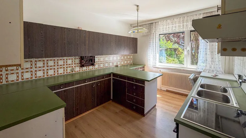 Ältere Küche mit grünen Arbeitsplatten, dunklen Holzschränken und Blick ins Grüne durch das Fenster.