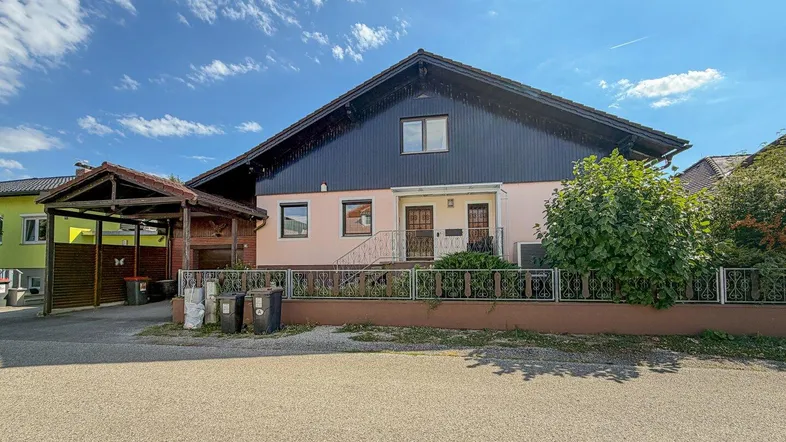 Hausfassade mit Carport und Vorgarten unter blauem Himmel.